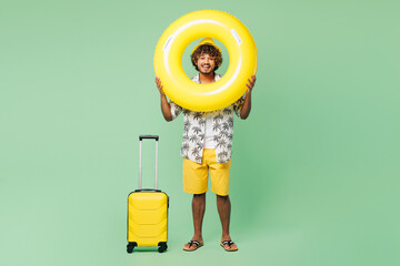 Traveler happy Indian man wear summer casual clothes hold suitcase bag rubber ring isolated on plain green color background. Tourist travel abroad in free time rest getaway. Air flight trip concept.