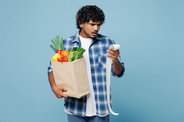 Wall Mural - Young sad mad Indian man he wear shirt hold brown craft bag for takeaway mock up with food products read cash bill receipt isolated on plain blue background. Delivery service from shop or restaurant.