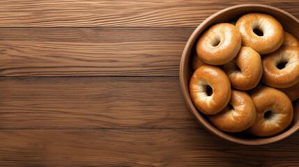 A bowl filled with a pile of plain bagels is set on a wooden table. The bagels have a shiny, golden-brown crust, indicating they are fresh and ready to eat.