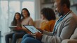 © napapat - A patient sitting comfortably on a couch at home, holding a tablet while talking to a doctor via telemedicine. In the background, family members provide emotional support, smiling and engaging