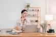 © kenchiro168 - A young woman sits at wooden desk, thoughtfully gazing at her laptop while surrounded by books and plants. serene atmosphere enhances her focus and creativity.