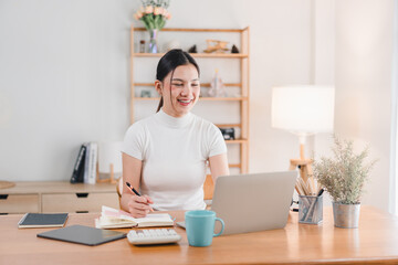 Wall Mural - A young woman is smiling while working on her laptop at cozy home office. She is taking notes and appears focused and happy in her bright, well organized workspace.