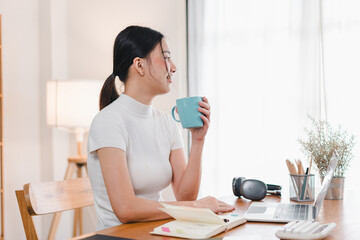 Wall Mural - A woman enjoys moment of relaxation while working at her desk, sipping from blue mug. cozy workspace features laptop, stationery, and warm ambiance.