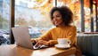 © FREDCassandra - Smiling woman working on laptop in cozy cafe with coffee, daytime, casual autumn attire, productivity and relaxation concept