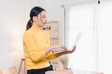 Wall Mural - A young woman in yellow sweater sits on table, working on her laptop in bright, airy room. scene conveys sense of productivity and comfort.