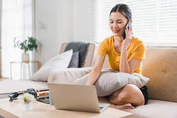 Wall Mural - A young woman is sitting comfortably on sofa, talking on her phone while working on laptop. She appears relaxed and engaged, surrounded by cozy living space.