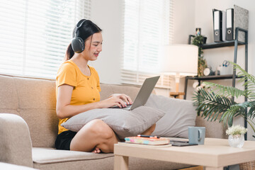 Wall Mural - A woman wearing headphones is sitting comfortably on sofa, working on her laptop with smile. cozy atmosphere is enhanced by natural light and stylish decor.