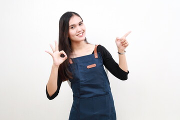 Young asian woman barista employee wear blue apron work in coffee shop pointing sideways while making okay hand gesture sign