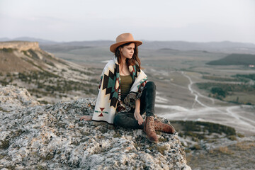 Wall Mural - Woman in hat and sweater sitting on mountain rock edge with boots, looking out into the distance