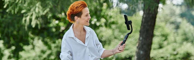 A redhead films video in park with selfie stick, smiling