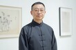 © Naru - A man with glasses, wearing a traditional Chinese black shirt, stands in a gallery setting with paintings in the background. He is looking at the camera with a calm expression. He appears to be an art