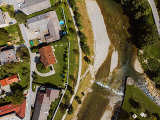 Aerial view of a residential area with houses, green lawns, and a river. The layout includes winding roads and a mix of buildings, showcasing a blend of nature and urban living.