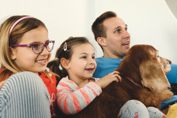 A family enjoying a cozy moment on the couch, watching TV. Two young girls, one with glasses, are smiling and laughing, while a man, presumably their father, looks surprised or amused. A dog is comfor