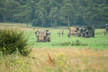 a squadron of British army Mastiff 2 6-wheel protected patrol vehicles and soldiers in a field, in action on a military exercise