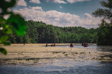 People are paddling down the river in canoes, with a forest visible in the distance with water lilies.