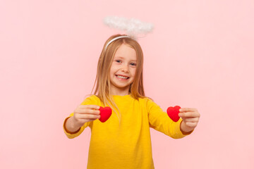 Wall Mural - Portrait of smiling delighted blonde little girl with nimb over head showing small hearts looking at camera with happiness, wearing yellow jumper. Indoor studio shot isolated on pink background.