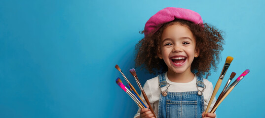 Wall Mural - A cheerful young girl holding paintbrushes and wearing an artist's beret, laughing against a blue background