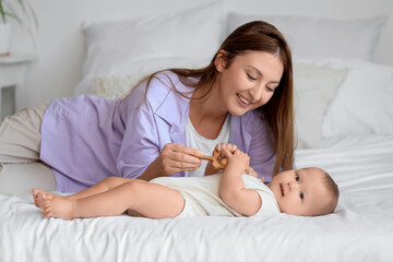 Beautiful young happy mother and cute little baby playing with rattle while lying in bedroom at home