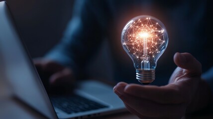 A person is holding a light bulb in their hand, with a laptop in the background