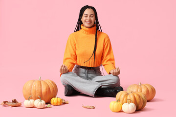 Beautiful young happy African-American woman with pumpkins and autumn leaves meditating on pink background