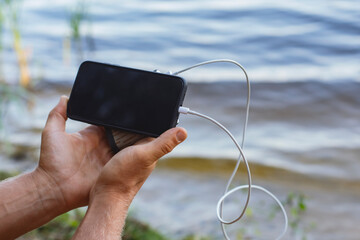 The guy is holding a portable charger with a smartphone in his hand. Man on a background of nature with a greenery and a lake