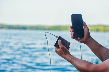 A man charges a smartphone with a power bank. The phone in hand is being charged with a portable charger against the backdrop of the lake.