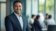 © Elmira - A confident Asian Indian businessman wearing a suit stands proudly with a smile in a sleek, modern office, surrounded by colleagues engaged in conversation