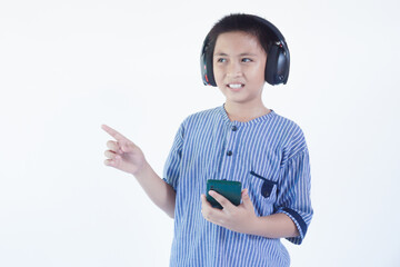 Smiling pretty Asian boy wearing headphones, holding phone, pointing up by finger, isolated