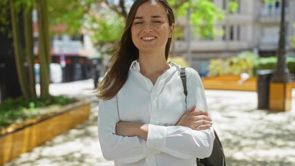 Wall Mural - A young blonde woman with crossed arms stands confidently in an outdoor city park, showcasing a serene urban setting with vibrant green trees and urban architecture in the background.