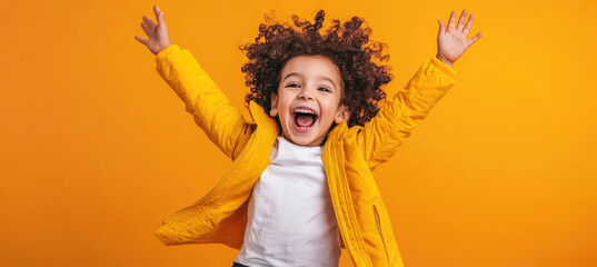 Wall Mural - A cheerful little girl with curly hair, wearing a white T-shirt and yellow jacket, jumping against an orange background