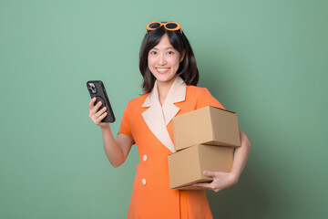 A happy woman in an orange dress holds two cardboard boxes and a phone against a green background. Perfect for themes of delivery, logistics, and e-commerce.