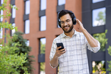 Satisfied and smiling young Indian man walking on a city street wearing headphones and using a mobile phone