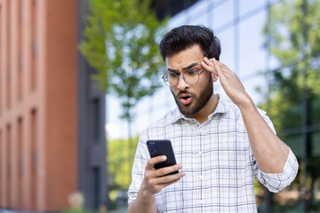 Wall Mural - A shocked young Muslim man stands outside a building, holding his head with his hand and looking at the screen of his mobile phone in surprise