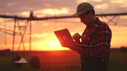 Wall Mural - silhouette agriculture. a male farmer works on a laptop in a field with green corn sprouts. corn is watered by irrigation machine. sunlight irrigation agriculture business concept