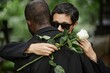 © Seventyfour - Medium shot of senior woman with white rose in hand giving comforting embrace to unrecognizable Black man grieving over loss of loved one at cemetery