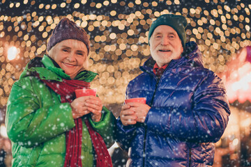 Wall Mural - Two people old married couple woman man enjoy x-mas time evening christmas atmosphere walk under newyear garland lights outdoors hold eggnog beverage mug