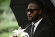 © Seventyfour - Portrait of adult Black man wearing sunglasses grieving loss of loved one at cemetery while hiding from rain under umbrella at memorial service, burial, copy space