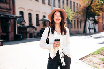 Lifestyle portrait of young stylish woman dressed in white blouse. Model walking with coffee cup in old town in sunny day. Beautiful smiling female in hat. Cheerful and happy. Takeaway coffee