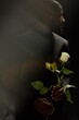 © Seventyfour - Vertical shot of African American gentleman in elegant suit holding white rose sadly looking away while grieving loss of loved one on black background in studio, ghost glare effect