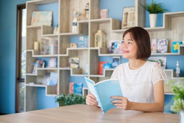 Woman in a bright room sitting at a desk reading a book near a large bookshelf