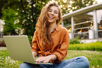 Wall Mural - A young woman enjoys a sunny afternoon in the park, engaging with her laptop while sitting comfortably on the grass surrounded by nature and flowers. Freelancer, Online education.