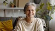 © VK Studio - A smiling older woman with short gray hair, wearing a cozy cream sweater, sitting in a comfortable, plant-filled living room.