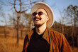 © maxbelchenko - Man wearing sunglasses and a large hat enjoys a sunny day outdoors in a grassy area surrounded by trees during autumn. Autumn scenery.