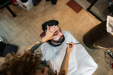 Professional barber carefully shaves and styles a man's beard using precise tools in a trendy urban salon, showcasing expertise and attention to detail in men's grooming.