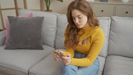 Canvas Print - A young attractive woman with blonde hair in a yellow sweater looks concerned while holding her phone, sitting in her living room at home.