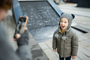 Canvas Print - Mother taking a photo of funny little boy having fun outdoors on autumn day. Kid walking in a city.