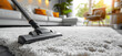 © Katerina Bond - Close-up of a vacuum cleaner head cleaning a plush grey carpet in a modern, sunlit living room with cozy furniture and decor, emphasizing cleanliness and home maintenance.