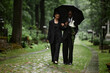 © Seventyfour - Full length shot of grieving multiethnic couple in black suits walking under umbrella along paved alley to visit grave and to place flowers at cemetery with lively greenery, copy space