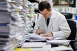 © Ilia - Doctor in a white coat sitting at a desk, filling out paperwork, with stacks of documents piled high around the desk. Concept of excessive paperwork in the medical field.