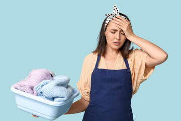 Poster - Portrait of tired young housewife in apron with dirty laundry on blue background